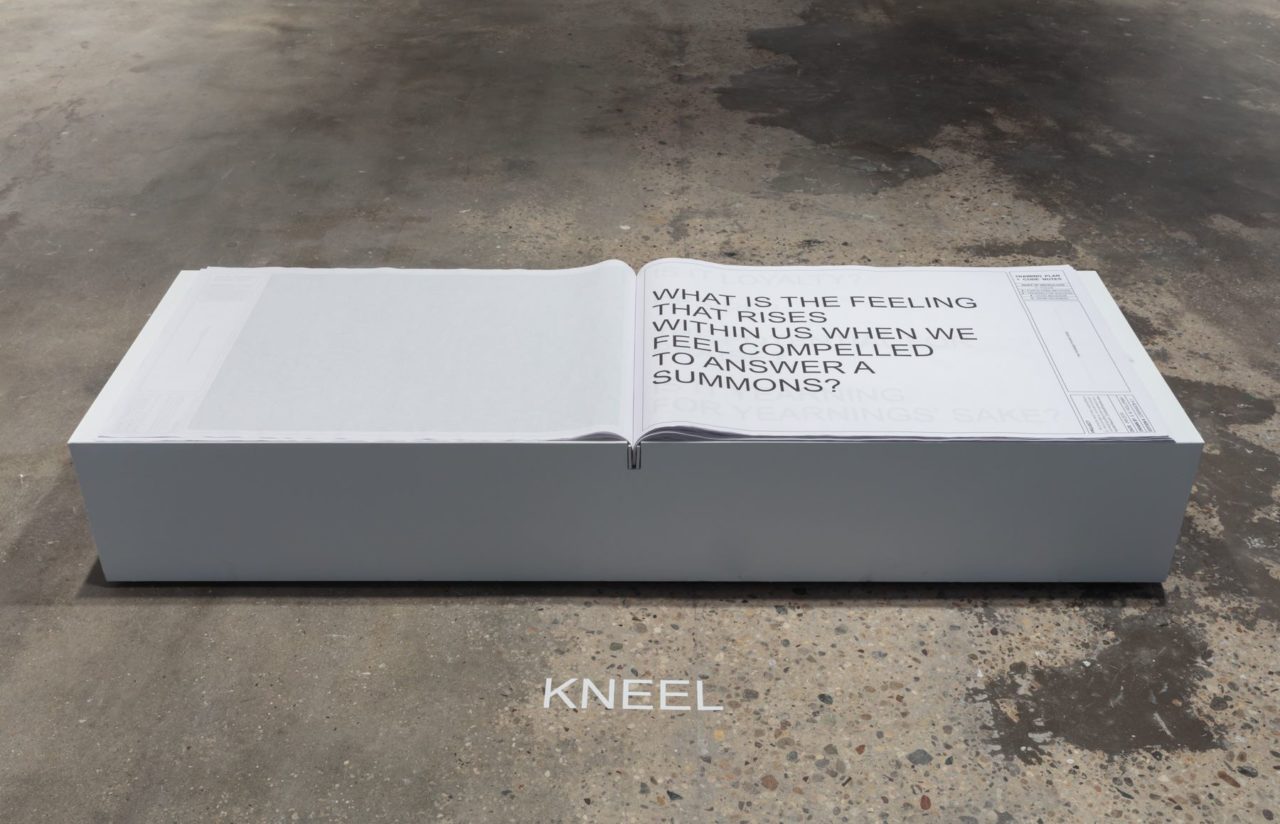 A large-format book opened with large text giving directions for puppy-play sits atop a white pedestal. Before the pedestal it commands the viewer to "kneel".