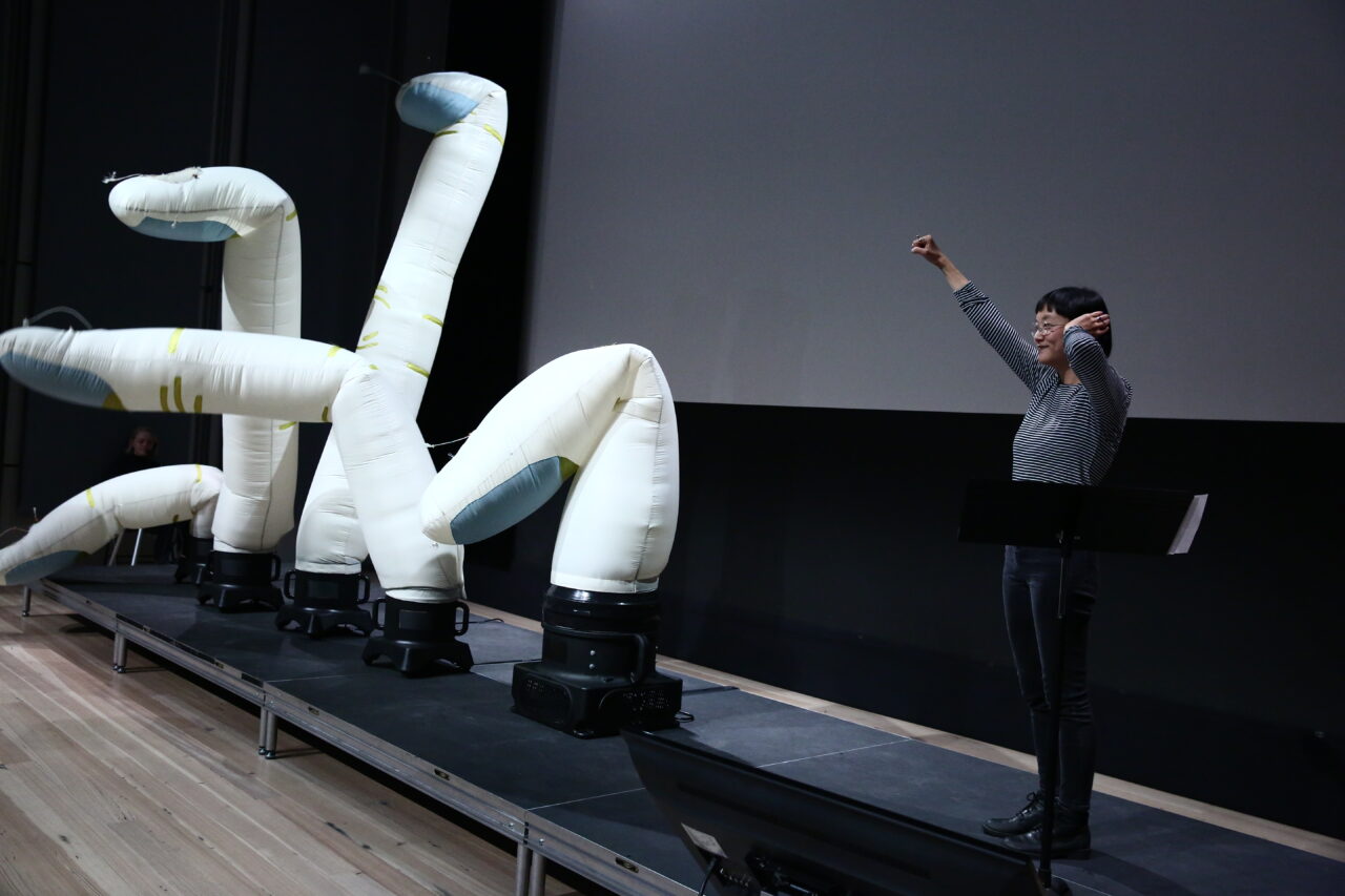 Christine Sun Kim performing on stage with what looks like four inflatable dancers.
