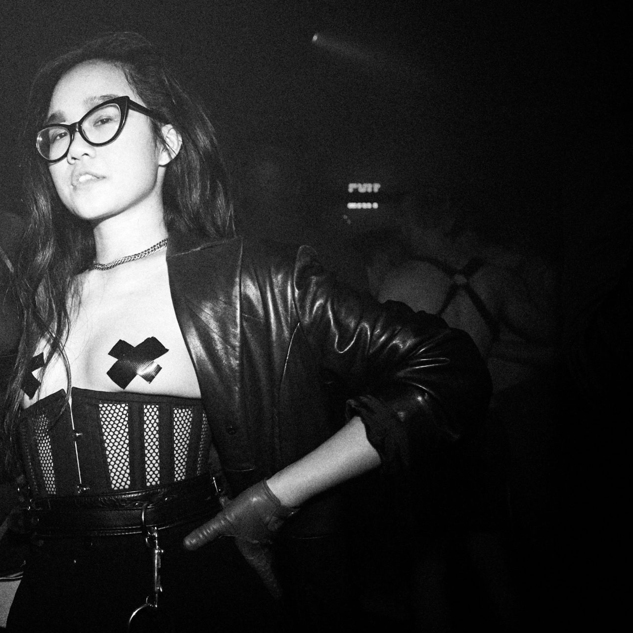 Black and white photo of a young Asian-American woman with her hand on her hip. She has long hair and is wearing cat-eye glasses, leather gloves, a leather blazer, and a mesh corset.