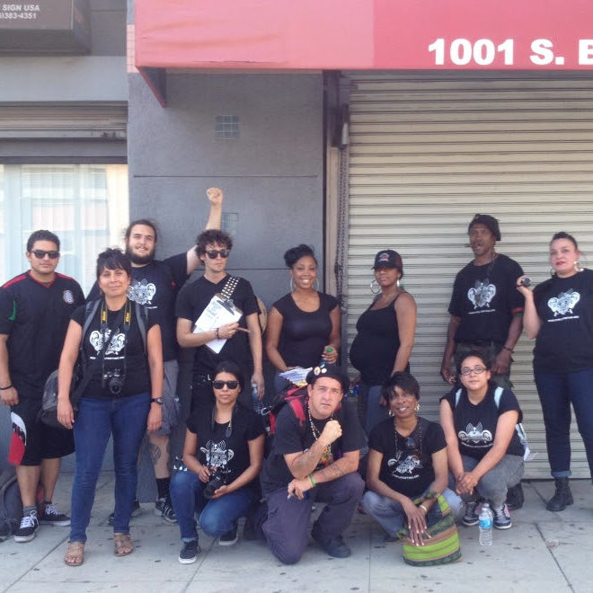 A predominantly BIPOC gathering of twelve adults smile as they stand and kneel on a sidewalk by a grey building in downtown Los Angeles. They are all dressed in black tops—many of which are a black t-shirt with a white screenprint of the Stop LAPD Spying Coalition logo—various dark-colored pants or shorts, and comfortable shoes. Four people are kneeling or squatting in the front row and eight people are standing in the back row. Some are wearing sunglasses, a hat, a backpack, or carrying a dSLR camera.