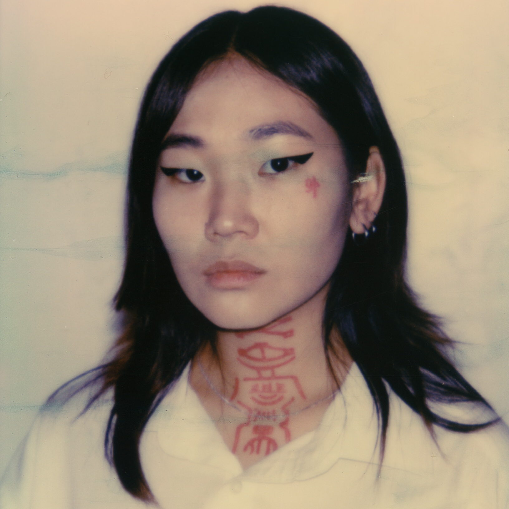 This image is of a young Korean woman of light/medium skin tone with dark hair and stark black eyeliner, she displays a red face and neck tattoo.