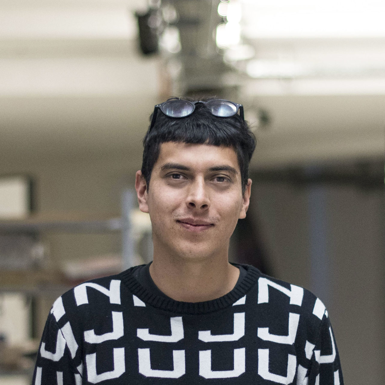 A light-brown Tropical South American Human slightly smiling with relatively big eyes and considerably big eye bags, glasses on his hair and a pullover that says JUAN multiple times. There’s a blurred background too - presumably at an art space in Berlin.