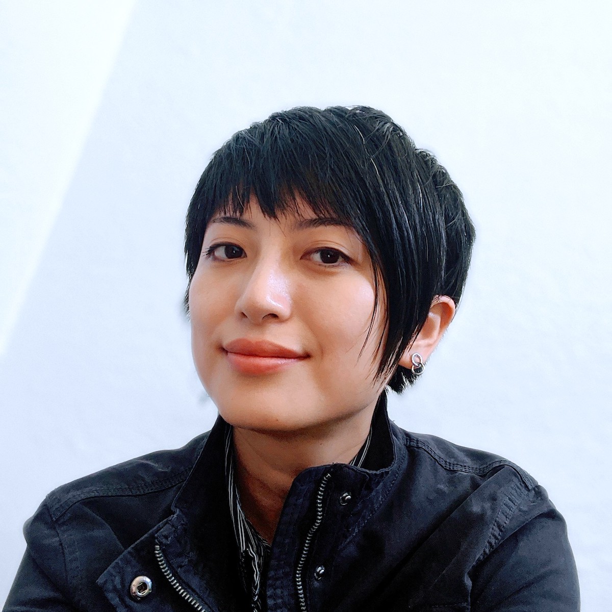 Photo of a non-binary Han Taiwanese descendent with a short, assymetrical haircut and a few strands of white hair. They have a diamond-shaped face, light-olive skin, almond-shaped eyes and a high nose bridge. They wear a black, unzipped jacket and a tiny earring consists of two intersecting rings. They appear to be sitting at the corner of the room and they look into the camera with a soft but confident expression.