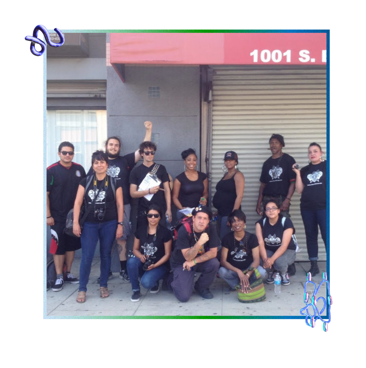 A predominantly BIPOC gathering of twelve adults smile as they stand and kneel on a sidewalk by a grey building in downtown Los Angeles. They are all dressed in black tops—many of which are a black t-shirt with a white screenprint of the Stop LAPD Spying Coalition logo—various dark-colored pants or shorts, and comfortable shoes. Four people are kneeling or squatting in the front row and eight people are standing in the back row. Some are wearing sunglasses, a hat, a backpack, or carrying a dSLR camera.