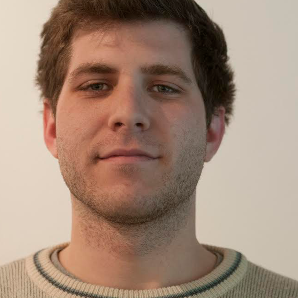 Light-skinned man in a beige sweater against an off-white background. They have light-brown hair, light stubble, pale grey eyes, and a very slight smile.