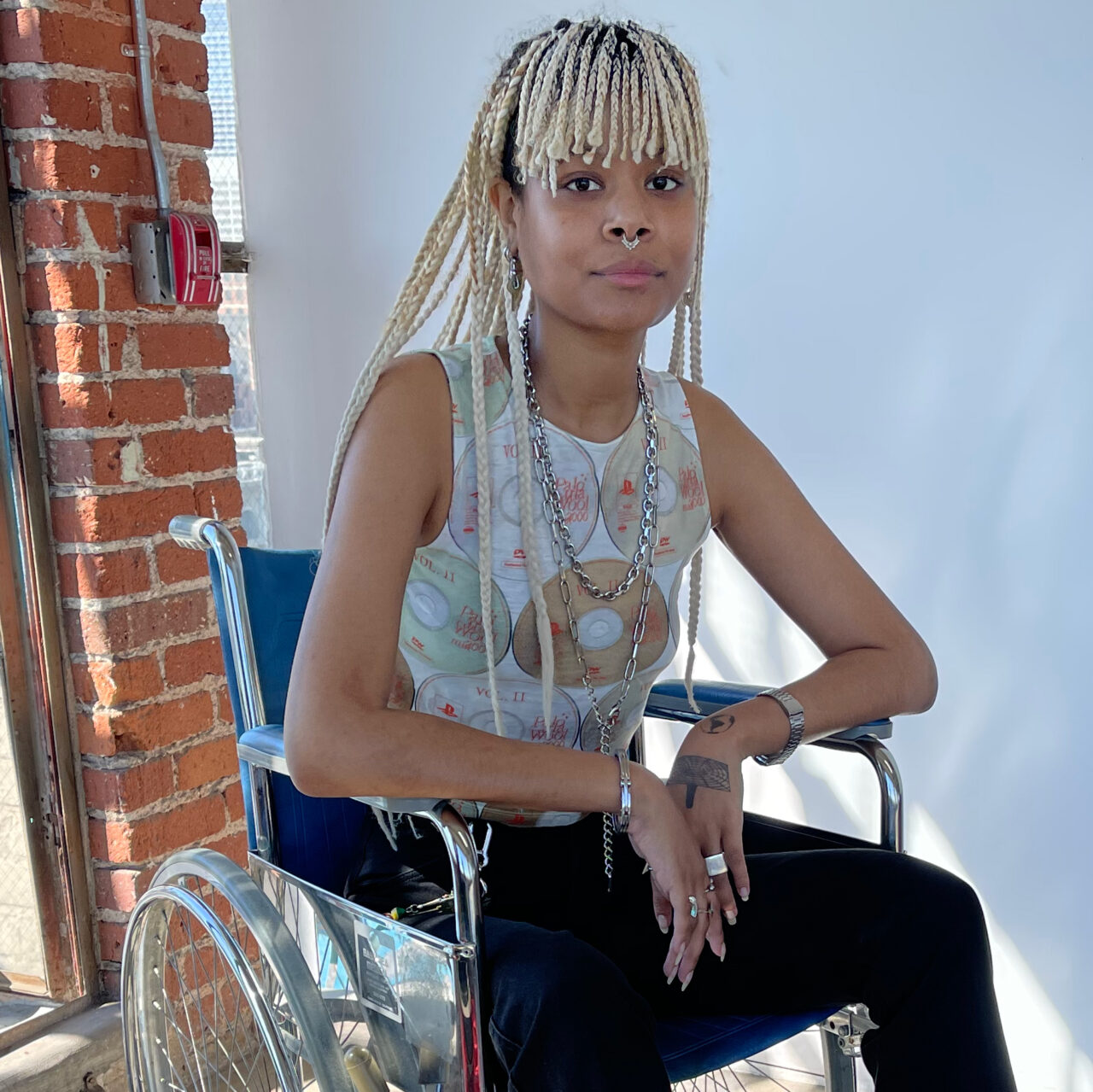 Panteha Abareshi sits in a blue leather-backed wheelchair. They are in a well-lit, brick and white-walled space, captured from the mid-bust upwards. They are wearing black pants, a sleeveless shirt printed with CDs and DVDs, silver chains around their neck, and silver rings on their fingers. Their hair is in blonde braids with bangs.