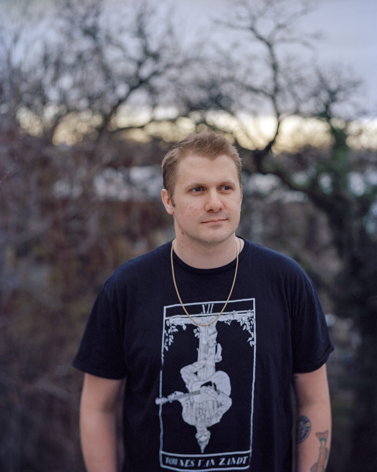 Brent, a white man with blonde hair, wearing a black T-shirt and gold chain, looks slightly past the camera, a slight smile on his lips.