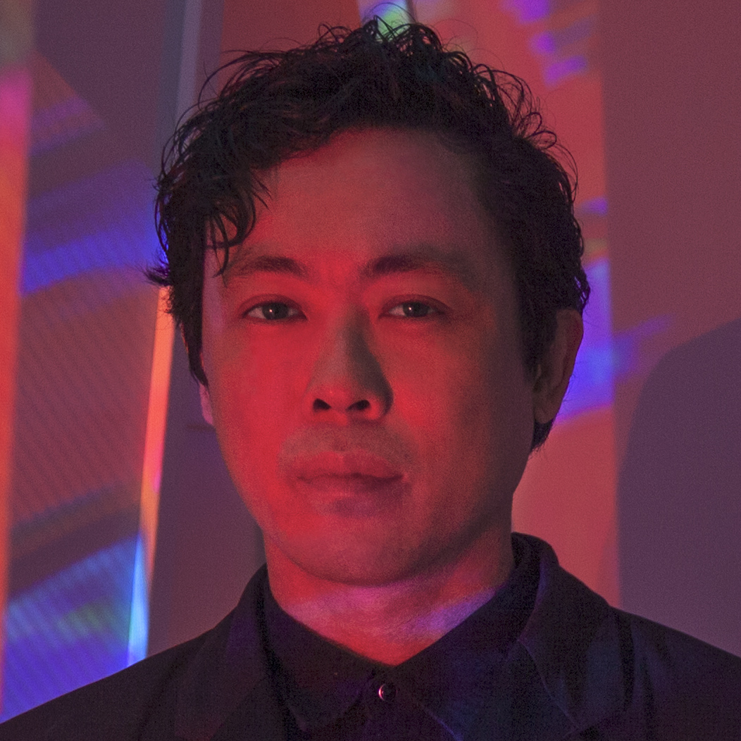 Peter, illuminated in red light against a red and blue background, looks directly at the camera. he has short, curly black hair and a black shirt.