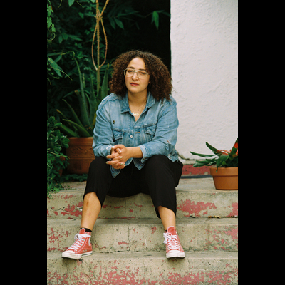 Zeina sits on a red brick staircase, with a white wall in the background. She has curly black medium-length hair, wears glasses, and a jean shirt with black pants.