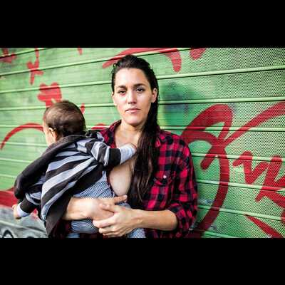 Daniela looks at the camera, her long brown hair swept back. She is holding a baby against her breast, its head turned away from the camera. she is wearing a red checkered shirt and the baby is wearing a blue and white striped sweater.