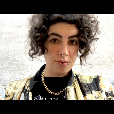 Nushin looks directly at the camera, eyebrows raised. She has three lip piercings, medium length curly-black hair, with a gold chain and a patterned yellow-and-black jacket.