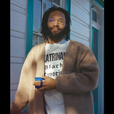 Ryan looks directly at the camera. He has long-ish dreadlocks and a black beard, is wearing a brown furry coat and a gold chain, and holding a blue mug.