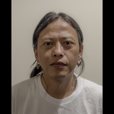 Subash looks directly at the camera, expressionless. He has long black-gray hair tied back in a ponytail and spiky silver earrings. He is wearing a white shirt.
